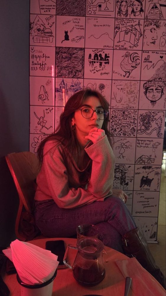 Yevgenya in glasses sitting in a café under red light, in front of a wall of hand-drawn doodle tiles.