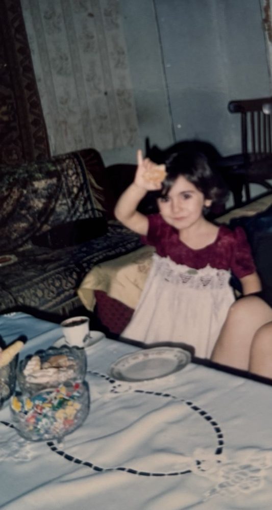 Childhood photo: a little girl in a red dress with a white lace front, waving at a family table with a lace tablecloth, damask throws and lace curtains behind her.